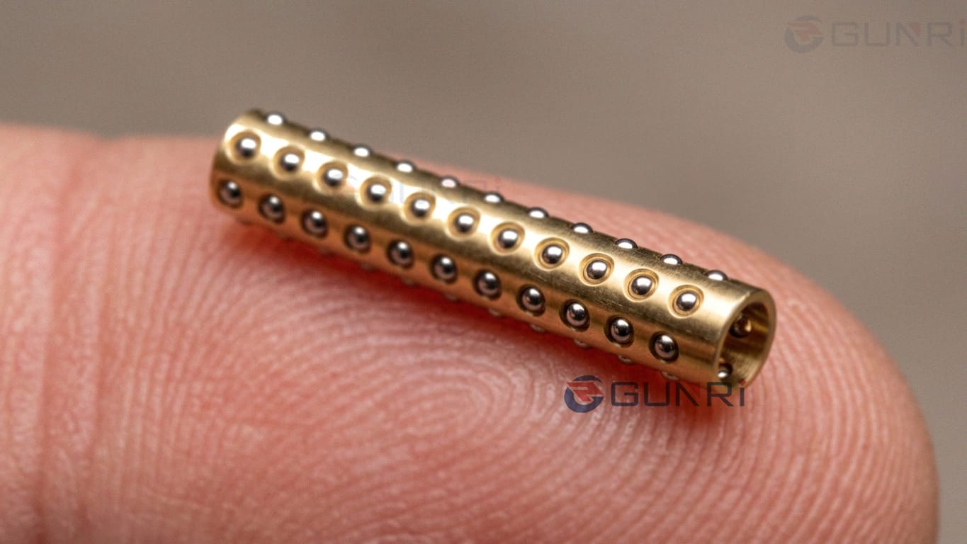 Miniature brass ball cage with precision steel balls held on a fingertip for size reference — custom miniature ball retainer for LVDT gage heads and precision linear motion